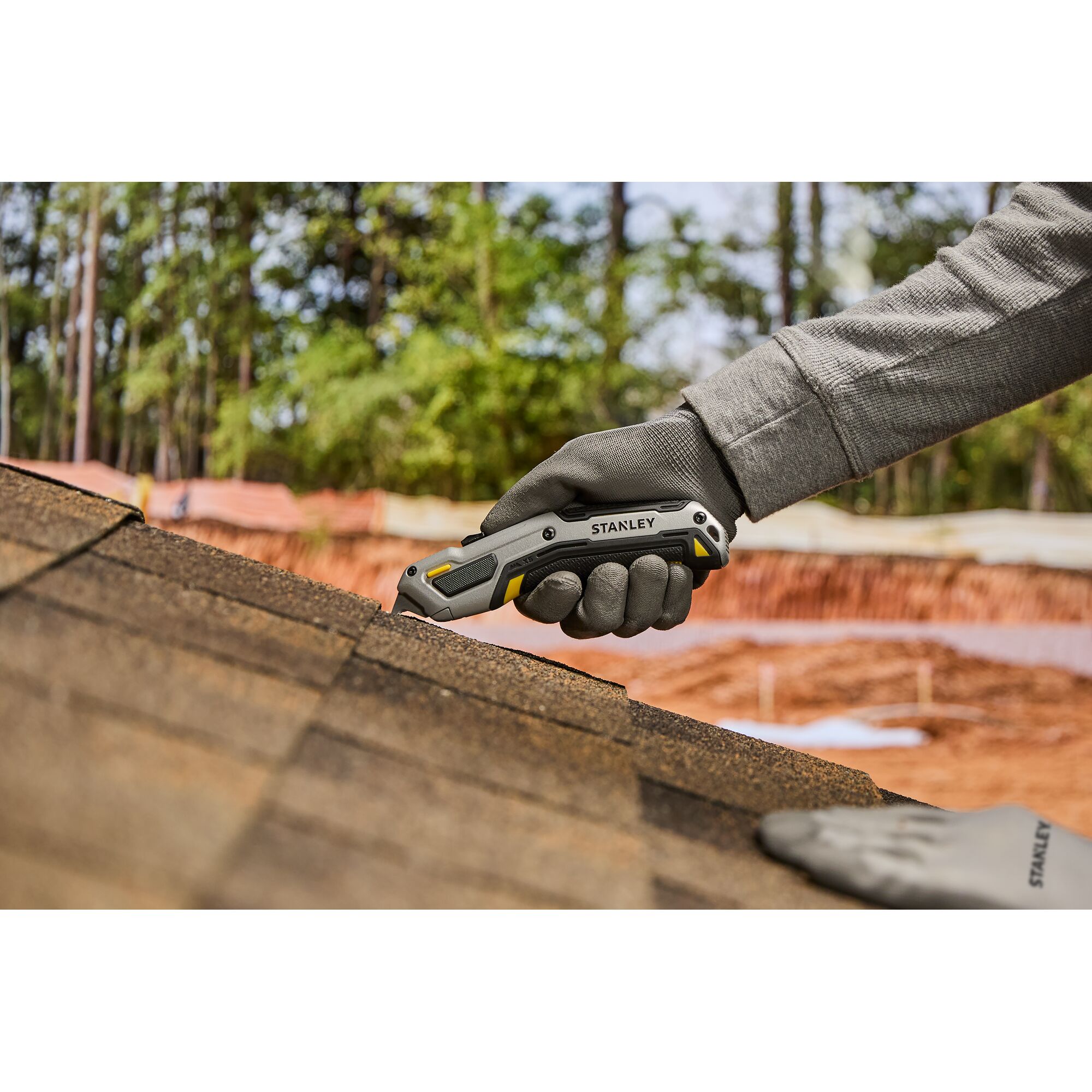 Une main gantée utilise un couteau utilitaire Stanley pour couper des bardeaux de toiture sur un toit, avec des arbres et des matériaux de construction en arrière-plan.